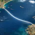 Aerial view of a glowing curved white line across the sea, connecting a rocky coastline to a coastal town, with several ships nearby.