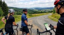 Three cyclists on a rural gravel road, one glancing back as the third rider checks a map on a phone mounted to a handlebar.