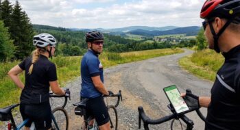 Three cyclists on a rural gravel road, one glancing back as the third rider checks a map on a phone mounted to a handlebar.