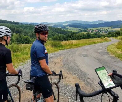 Three cyclists on a rural gravel road, one glancing back as the third rider checks a map on a phone mounted to a handlebar.