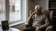 Older man sits in a beige armchair by a window, hand to chin, deep in thought with a framed photo on the table nearby