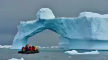 Group of people in an inflatable boat wearing red life jackets near a blue iceberg arch in calm water.