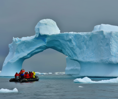 Group of people in an inflatable boat wearing red life jackets near a blue iceberg arch in calm water.