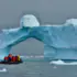Group of people in an inflatable boat wearing red life jackets near a blue iceberg arch in calm water.