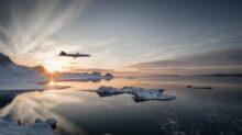 Jet plane flying low over a snowy coastline at sunset with icy mountains in the background and a calm reflective inlet below.