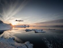 Jet plane flying low over a snowy coastline at sunset with icy mountains in the background and a calm reflective inlet below.