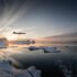 Jet plane flying low over a snowy coastline at sunset with icy mountains in the background and a calm reflective inlet below.