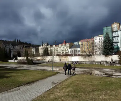 Four people walk away on a brick path through a park toward a row of pastel historic buildings under a dark stormy sky.