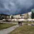 Four people walk away on a brick path through a park toward a row of pastel historic buildings under a dark stormy sky.