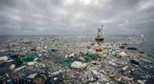Ocean surface littered with plastic bottles, bags, nets and debris under a cloudy sky, with a small platform in the distance