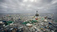 Ocean surface littered with plastic bottles, bags, nets and debris under a cloudy sky, with a small platform in the distance