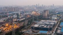 Aerial view of an urban industrial complex with cooling towers, smokestacks, rail yards, and power lines at dusk.