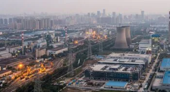 Aerial view of an urban industrial complex with cooling towers, smokestacks, rail yards, and power lines at dusk.