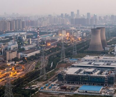 Aerial view of an urban industrial complex with cooling towers, smokestacks, rail yards, and power lines at dusk.
