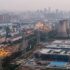 Aerial view of an urban industrial complex with cooling towers, smokestacks, rail yards, and power lines at dusk.