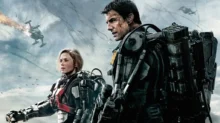 Two soldiers in bulky powered exoskeletons stand on a smoky battlefield, with helicopters in the sky behind them.