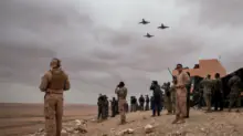 Soldiers in desert terrain watching four jets fly overhead near a military camp.