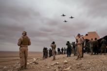 Soldiers in desert terrain watching four jets fly overhead near a military camp.