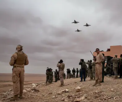 Soldiers in desert terrain watching four jets fly overhead near a military camp.