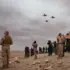 Soldiers in desert terrain watching four jets fly overhead near a military camp.