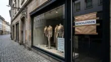 Storefront window displaying three beige suits on mannequins; taped 'GESCHLOSSEN' sign indicates the shop is closed.