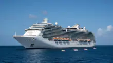 White cruise ship anchored on a calm blue sea under a clear sky, with orange lifeboats visible along its side.