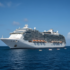 White cruise ship anchored on a calm blue sea under a clear sky, with orange lifeboats visible along its side.