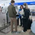 Na letišti stojí cestující společně se zaměstnankyní letiště, která jim pomáhá s odbavením